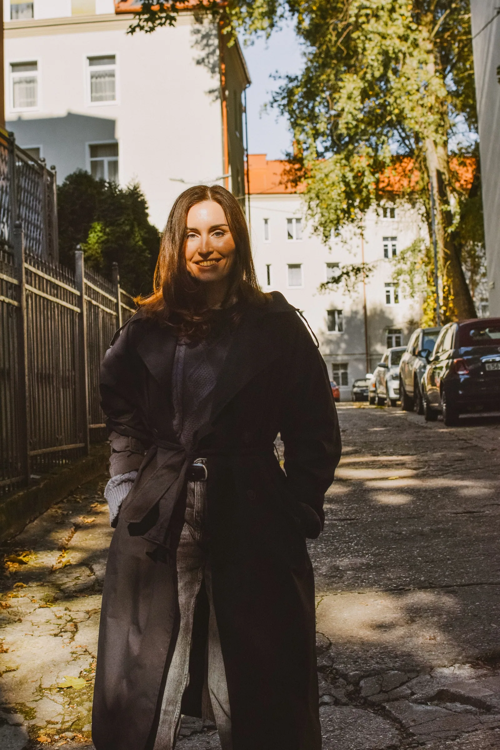Длинное чёрное пальто, руки в карманах. Солнечная улица, тени от деревьев на асфальте, и она улыбается прямо в объектив