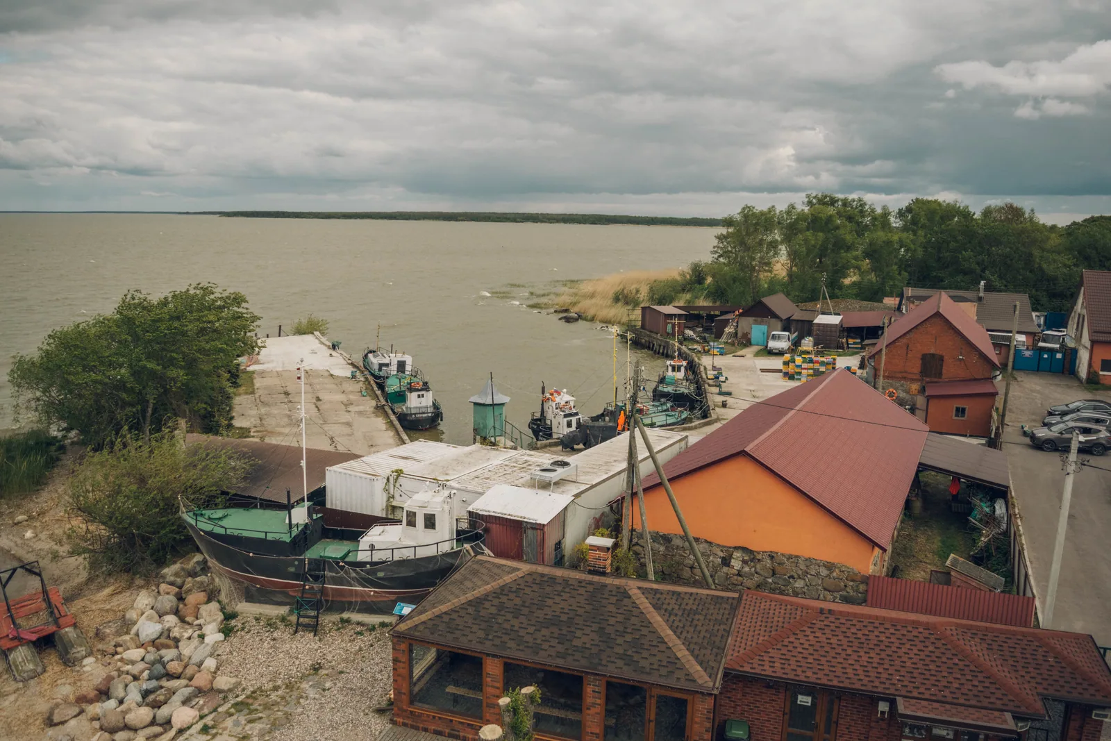Сверху видно, как посёлок повернулся к воде: причал, катера, красные крыши сараев и тростник у берега
